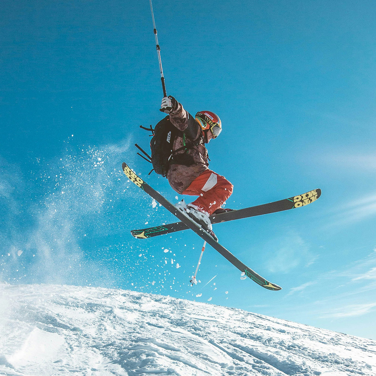 Skifahrer in roter Skihose und brauner Jacke beim Sprung in der Luft, blauer Himmel im Hintergrund