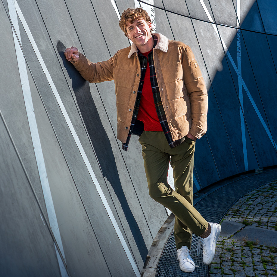 Mann in beiger Winterjacke mit Fellkragen, roter Strickpullover, olivgrüne Hose, draußen angelehnt
