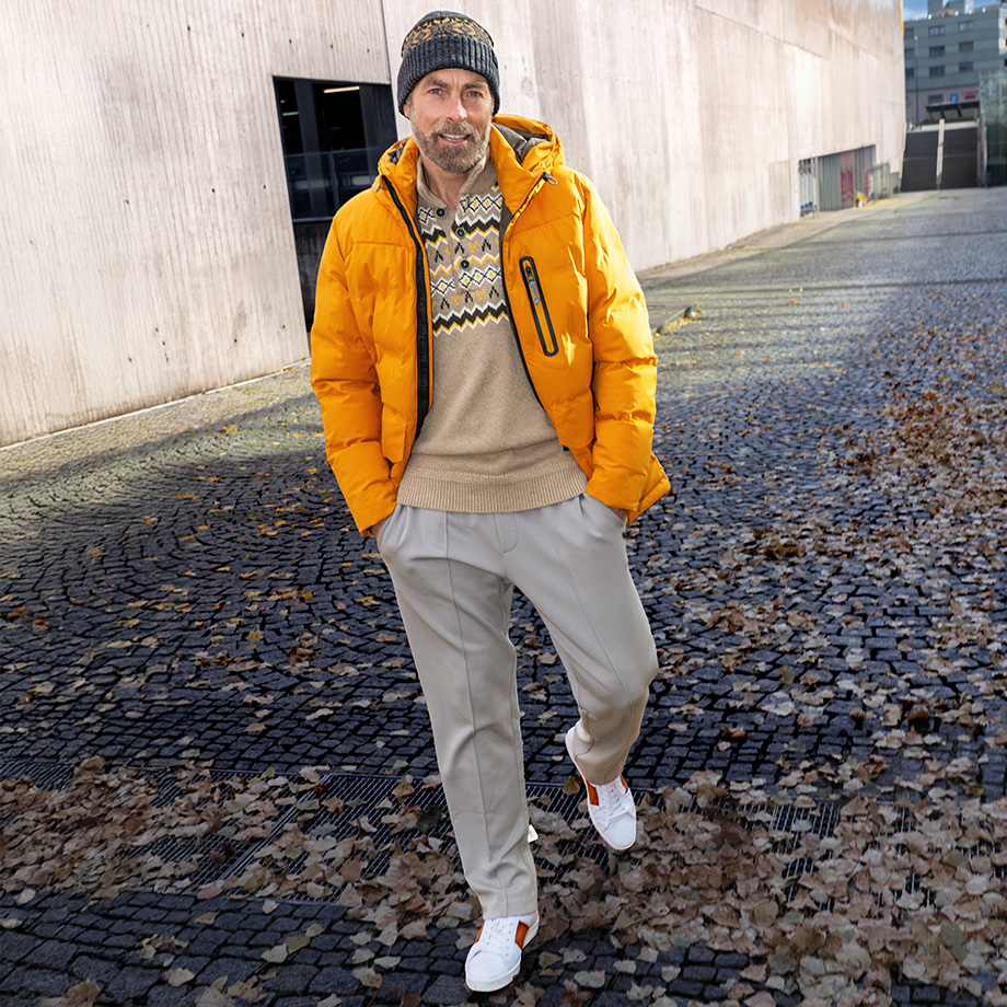 Mann in orangefarbener Steppjacke und beigem Pullover mit Zopfmuster, graue Hose und Mütze