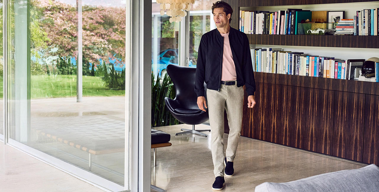 Ein Mann trägt einen dunkelblauen Blouson über einem rosa T-Shirt, kombiniert mit einer grauen Hose von Alberto und dunkelblauen Slippern. Er steht in einem modernen Haus mit großzügigen Glasfronten, die für eine helle und offene Atmosphäre sorgen.