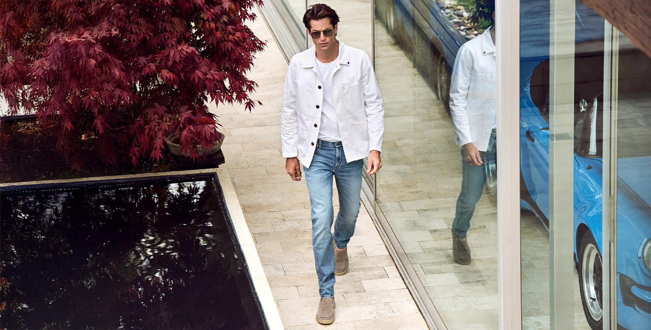 Ein modisch gekleideter Mann mit weißem Overshirt und blauer Jeans spaziert draußen entlang einer Glasfassade.