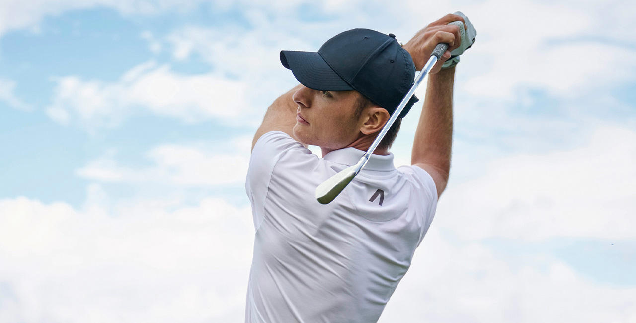 Ein Mann in weißen Golf-Polo & blauer Cap holt zum Abschlag aus. Im Hintergrund ist weiß blauer Himmel.