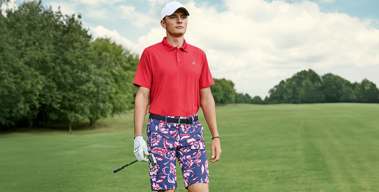Ein Mann läuft über einen weitläufigen Golfplatz. Er trägt ein rotes Polo-Shirt und dazu eine dunkelblau-rot gemusterte Golfshort. In der rechten Hand hält er einen Golfschläger.