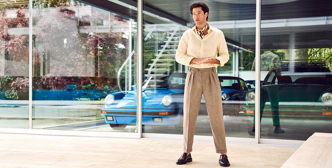 Ein Mann trägt eine locker geschnittene beige Bundfaltenhose, ein beiges Hemd und ein Nicki Tuch. Er steht in einem modernen Gebäude mit großer Glasfront.   Ein Mann trägt eine locker geschnittene, beige Bundfaltenhose, ein hellbeiges Hemd und ein elegantes Nickituch. Er steht in einem modernen Gebäude mit einer großen Glasfront.