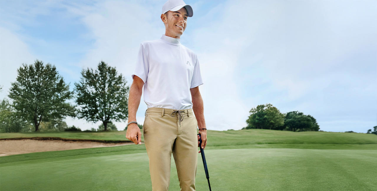 Ein Mann auf einem Golfplatz. Er trägt ein weißes Polo-Shirt, eine beige Hose und eine weiße Golfkappe. In der linken Hand trägt er einen Golfschläger.