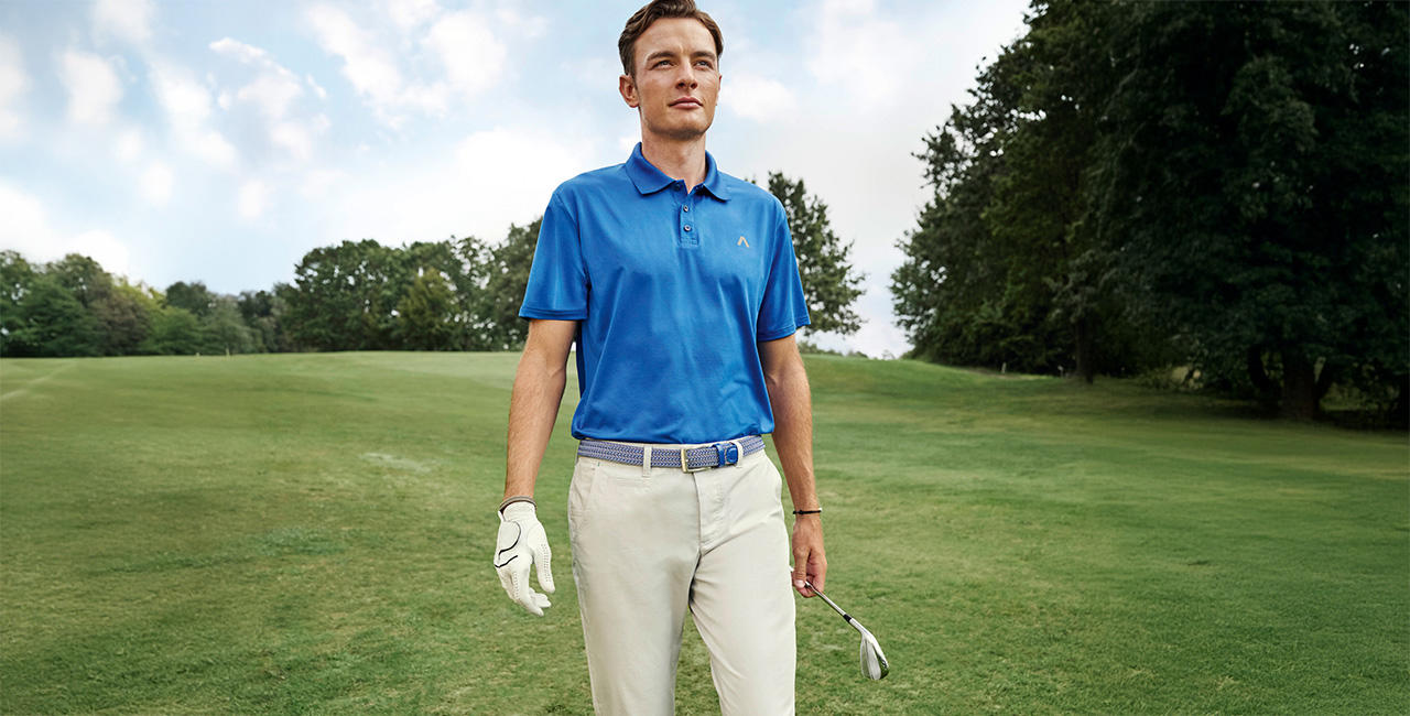 Ein junger Mann in einem blauen Polohemd und beigen Hosen steht auf dem Golfplatz mit Handschuhen und Golfschlägern.