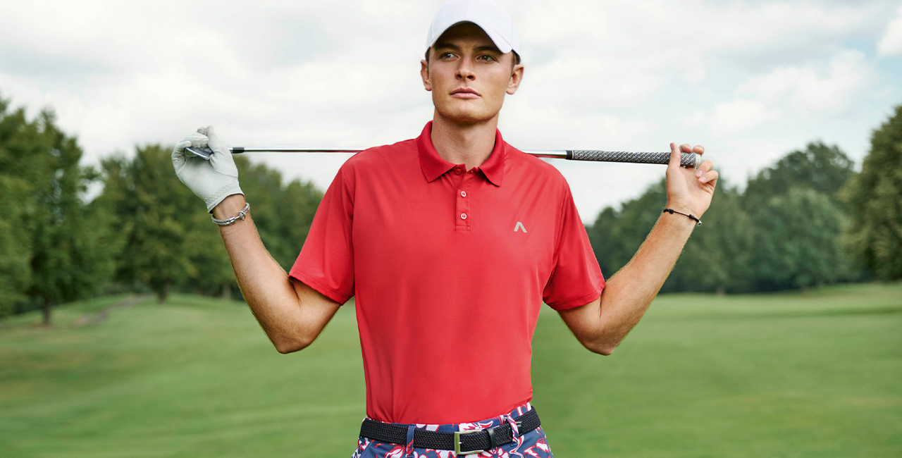 Ein Mann steht auf einem weitläufigen Golfplatz. Er trägt ein rotes Polo und weißes Cap. Hinter den Schultern trägt er einen Golfschläger.