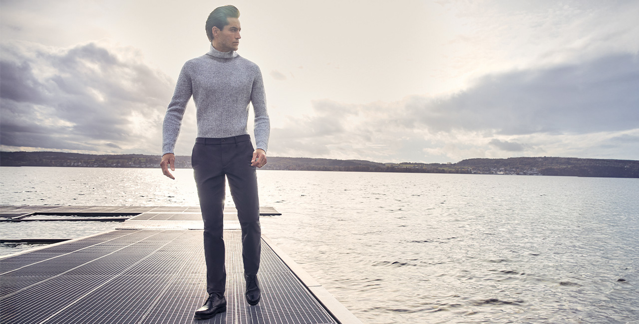 Ein Mann steht am Steg eines Sees. Er trägt einen hellgrauen Rollkragenpullover und dunkelgraue Hosen.
