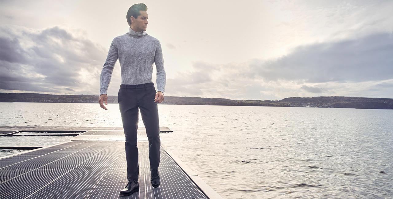 Ein Mann steht am Steg eines Sees. Er trägt einen hellgrauen Rollkragenpullover und dunkelgraue Hosen.