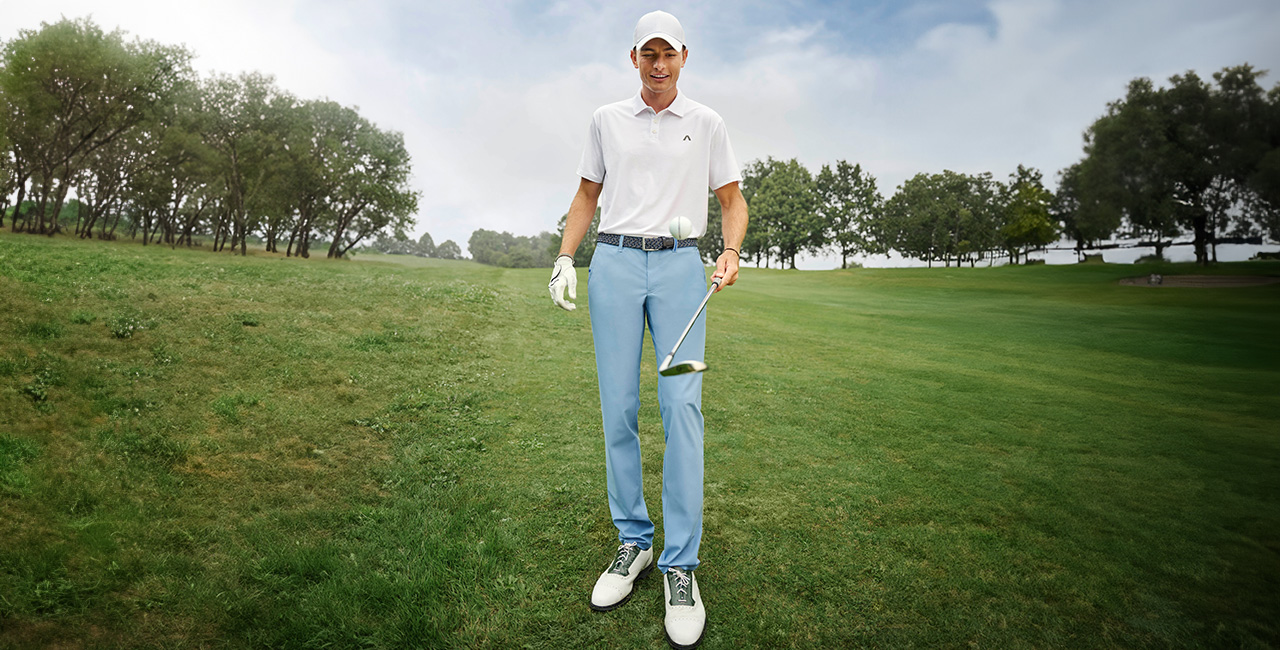Ein Mann steht auf einem weitläufigen Golfplatz. Er trägt ein weißes Polo, hellblaue Golfhosen und ein weißes Cap.