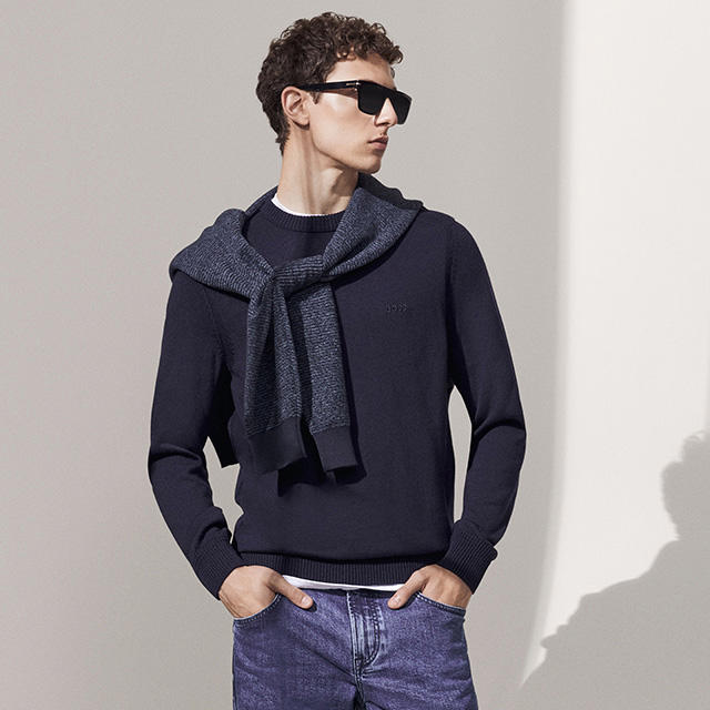 Ein Model mit Sonnenbrille trägt einen dunkelblauen Pullover, über der Schulter hat er einen weiteren blauen Pullover lässig gebunden. Dazu trägt er eine Jeans.