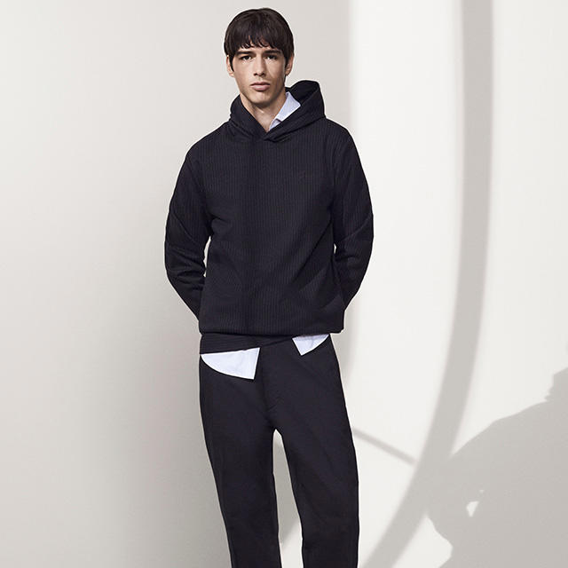 Ein Mann trägt eine schwarze Hose und einen schwarzen Hoodie mit einem weißen Hemd darunter.