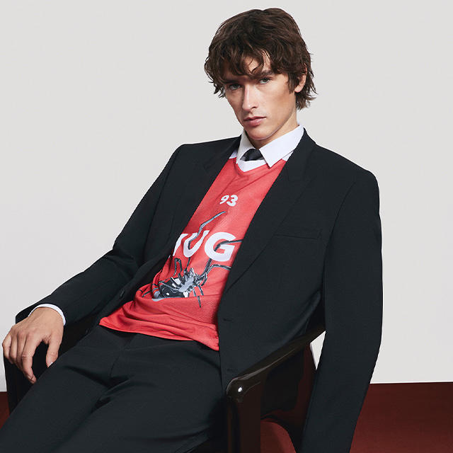 Ein Model trägt ein schwarzes Sakko über einem roten Shirt mit weißer Aufschrift und sitzt entspannt auf einem Stuh