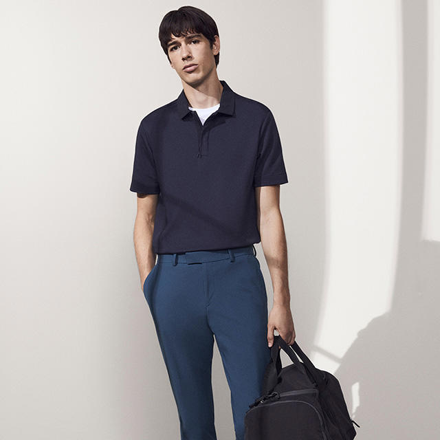 Ein junger Mann trägt ein dunkelblaues Poloshirt und eine blaue Hose, während er eine schwarze Tasche hält.