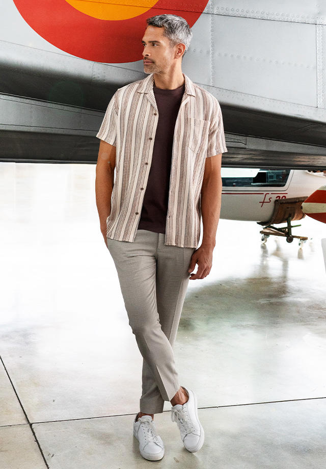 Ein Mann trägt ein gestreiftes Kurzarm-Hemd und eine beige Hose, während er in einer Flugzeughalle steht.