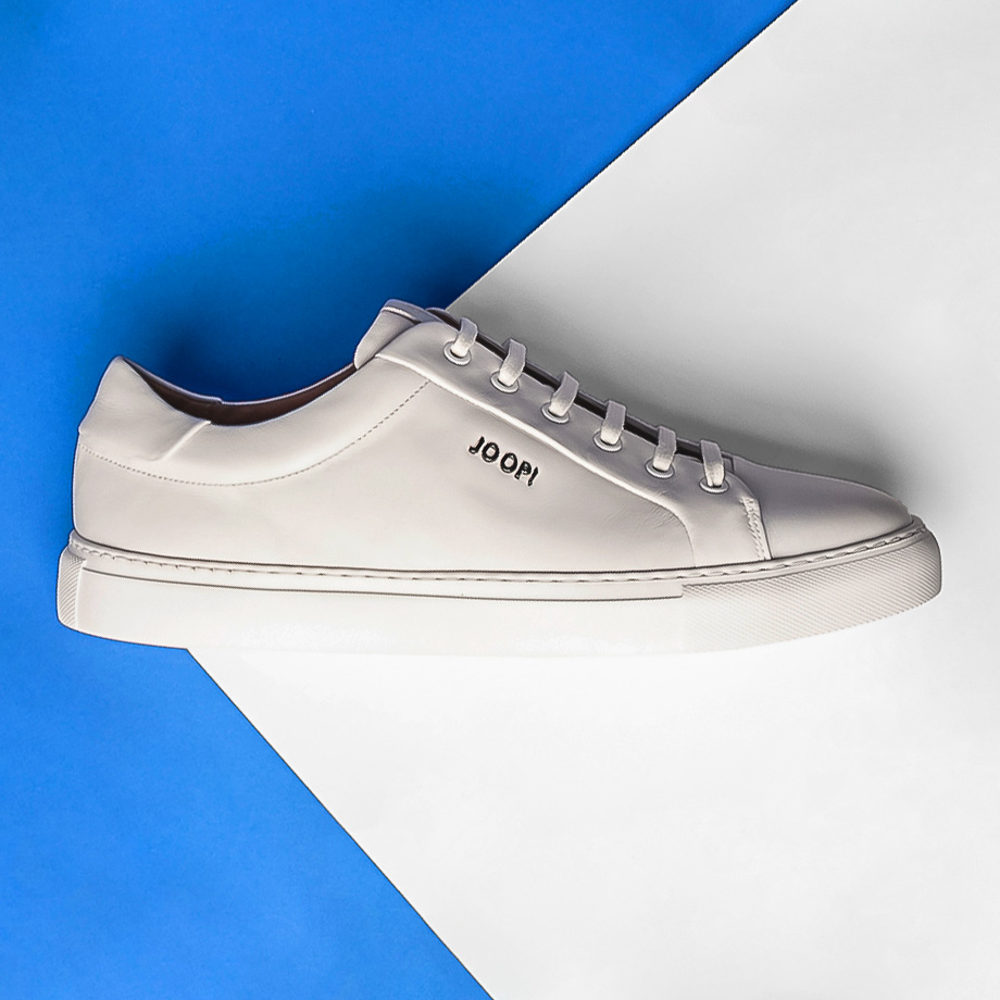 Ein weißer Sneaker von JOOP! liegt auf einem blauen und weißen Hintergrund.
