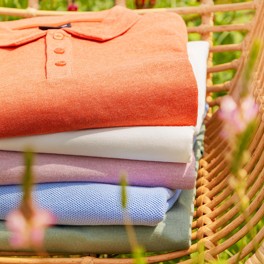Ein Stapel bunter Poloshirts in Orange, Weiß, Lila und Blau liegt auf einem Stuhl mit Blumen im Vordergrund.
