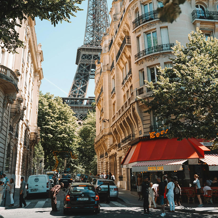 Eine belebte Pariser Straße mit klassischen Haussmann-Gebäuden, Bäumen und dem Eiffelturm im Hintergrund. Menschen schlendern über den Bürgersteig, einige sitzen in einem Café mit roter Markise, während Autos und Motorroller die Straße entlangfahren. Die Szene wird von sommerlichem Sonnenlicht beleuchtet.