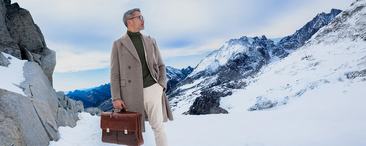 Ein Mann steht vor einer verschneiten Berglandschaft und schaut zur Seite. Er trägt eine Brille, einen beigen Mantel und einen braunen Rollkragenpulli.