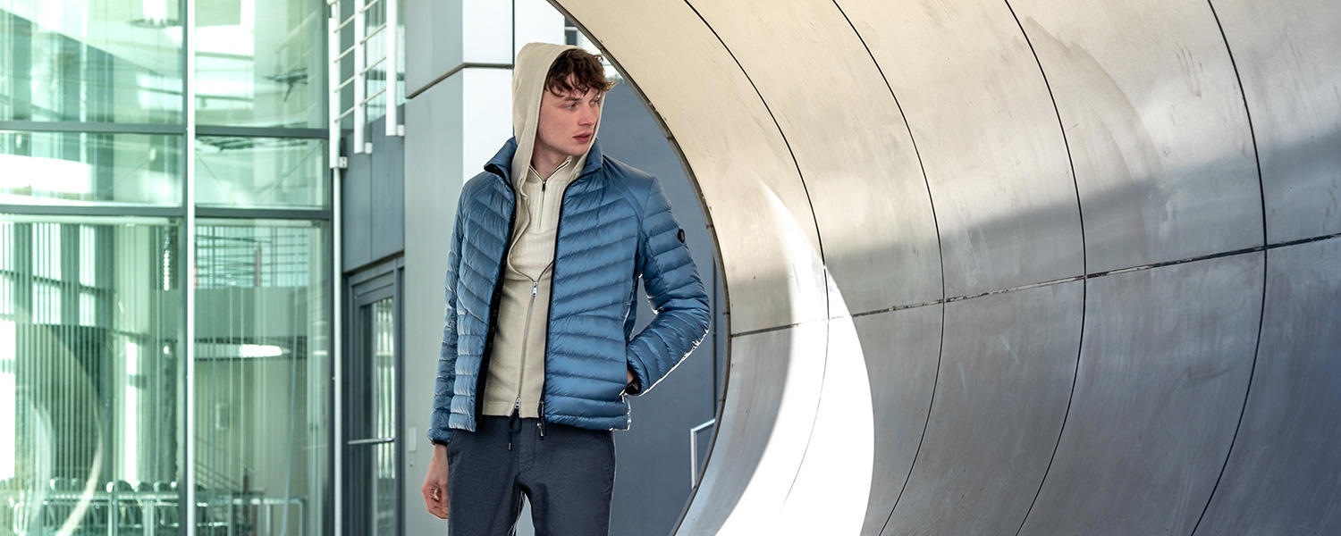 Ein junger Mann in einem Layering-Look aus Strick-Troyer, Strickjacke mit Kapuze und blauer Steppjacke steht in einer urbanen Umgebung.