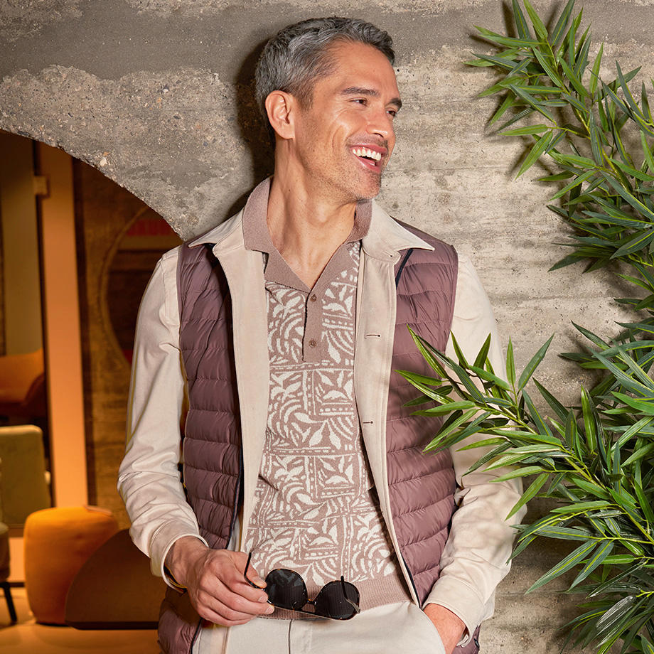 Ein Mann mit Sonnenbrille in der Hand hat ein beige gemustertes Strickpolo an, darüber ein beiges Overshirt und eine braune Steppweste. Er steht inmitten einer warmen Atmosphäre vor eine Betonwand 