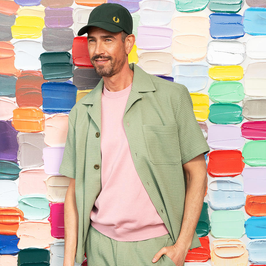 Ein Mann mit einem rosa Strickshirt und einem grünen Kurzarmhemd darüber steht vor einer bunten, künstlerischen Wand