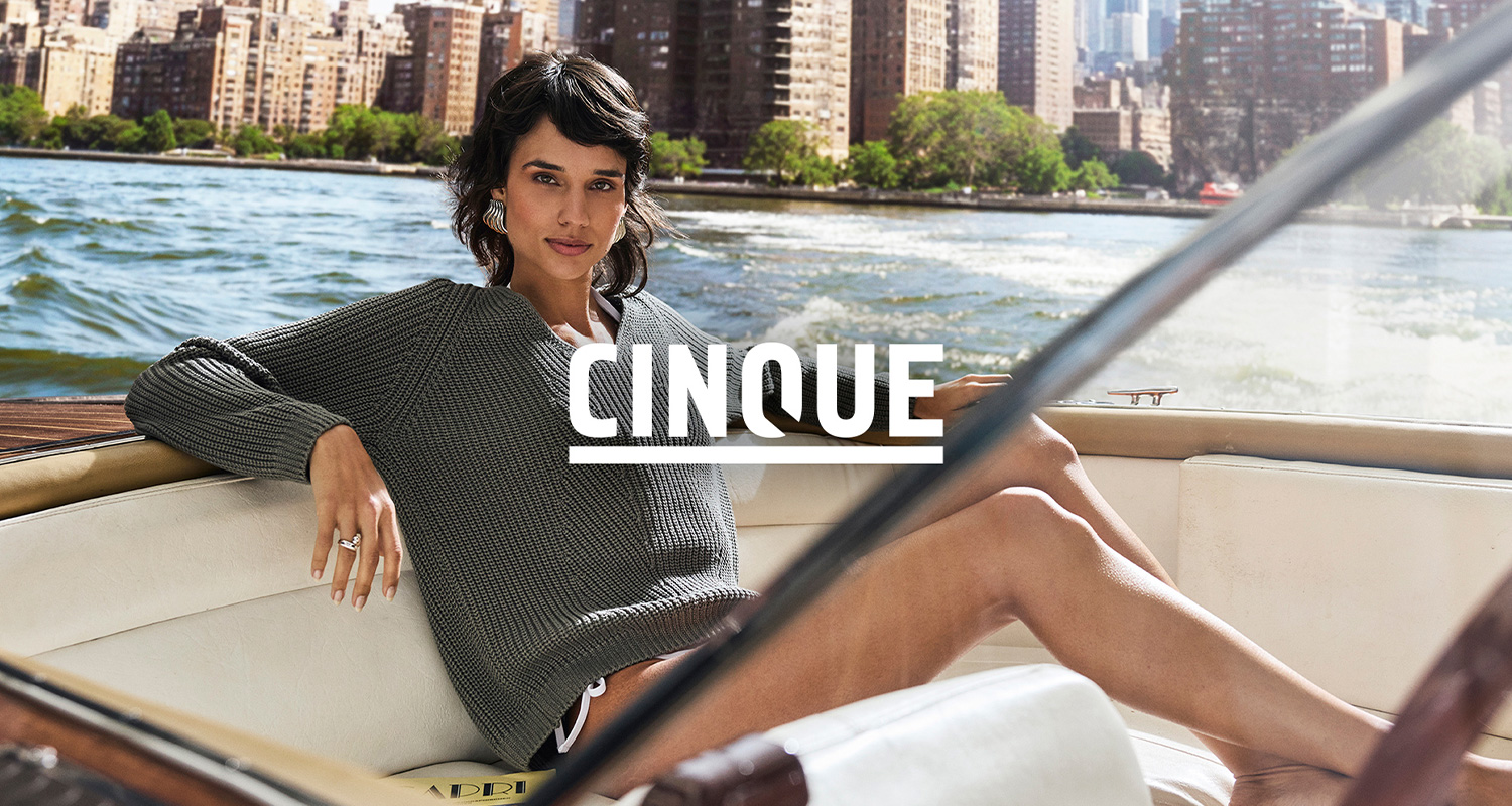 Eine Frau in einem grauen Pullover sitzt entspannt auf einem Boot, mit einer Wasserlandschaft und Stadtgebäuden im Hintergrund. Logo: CINQUE.