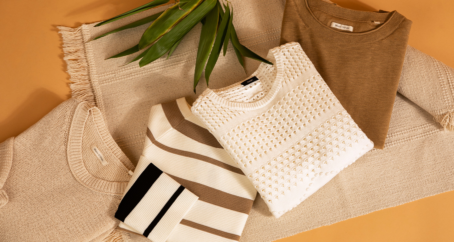 Flach gelegte Strickmode in natürlichen Farben, darunter beige und weiße Pullover, auf einem Stoffuntergrund mit grünen Blättern als Dekoration.