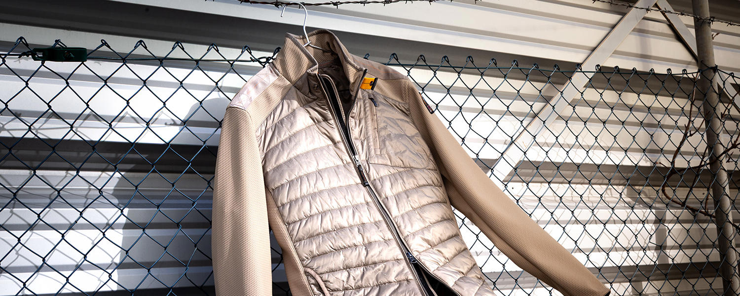 Eine Jacke im Materialmix hängt an einem Stacheldraht vor einer grauen Wand und einem Zaun. Einige Regentropfen glitzern auf der Jackenoberfläche