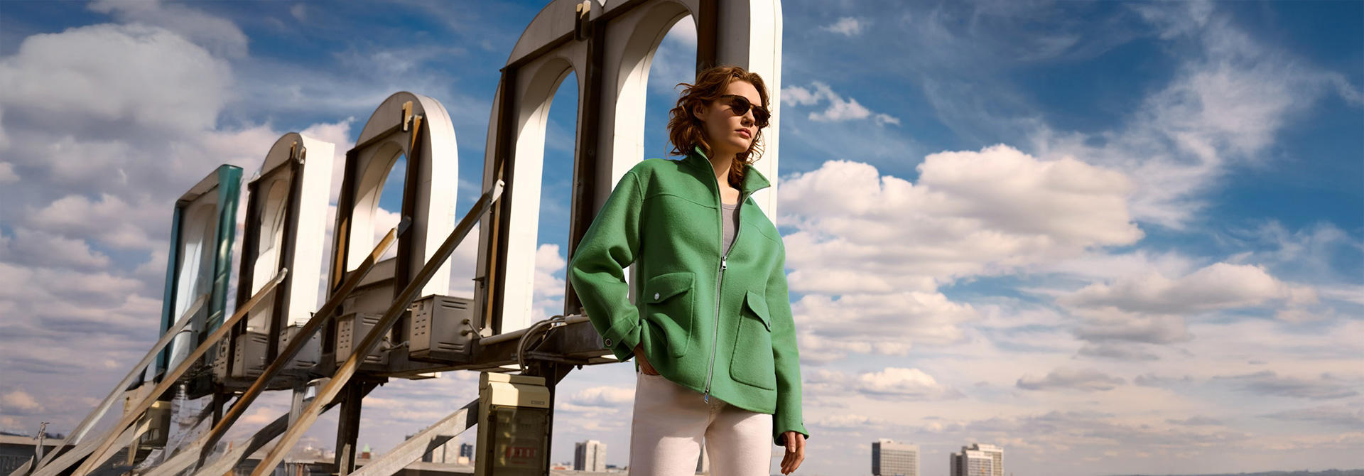 Eine Dame trägt eine grüne Wolljacke und eine Sonnenbrille. Sie steht auf einem Dach vor einem großen Werbeschild.