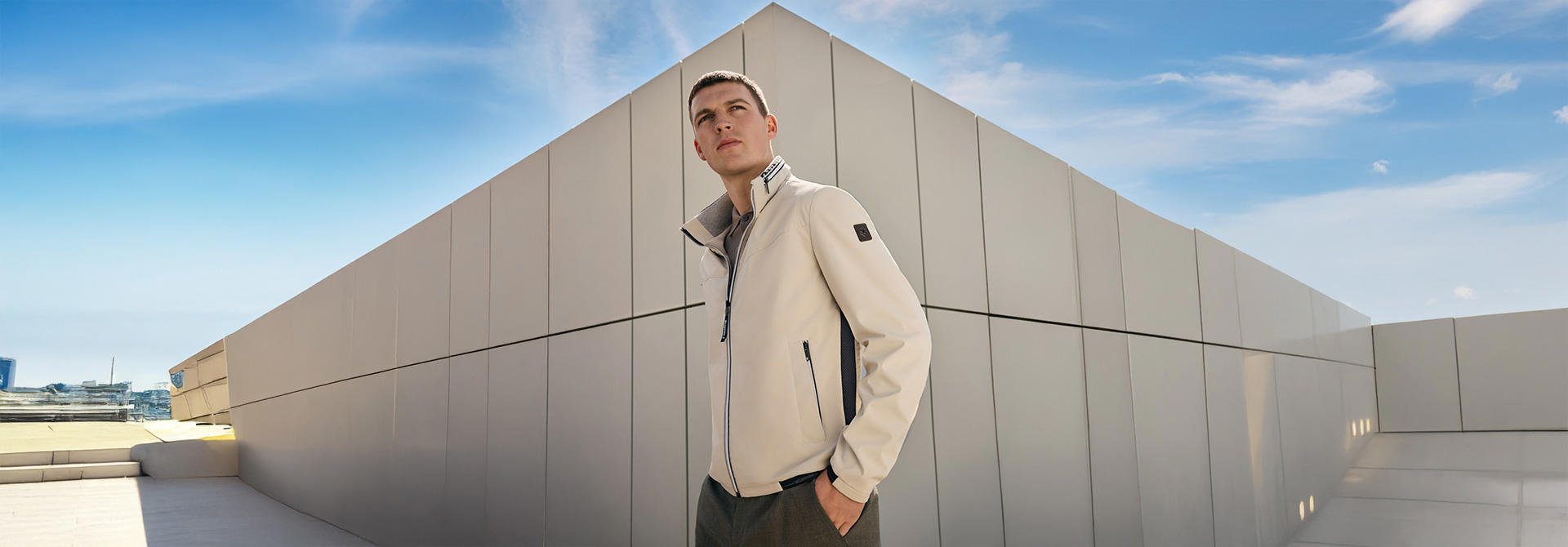 Ein Mann in einer hellbeigen Funktionsjacke steht vor einem grauen Gebäude.