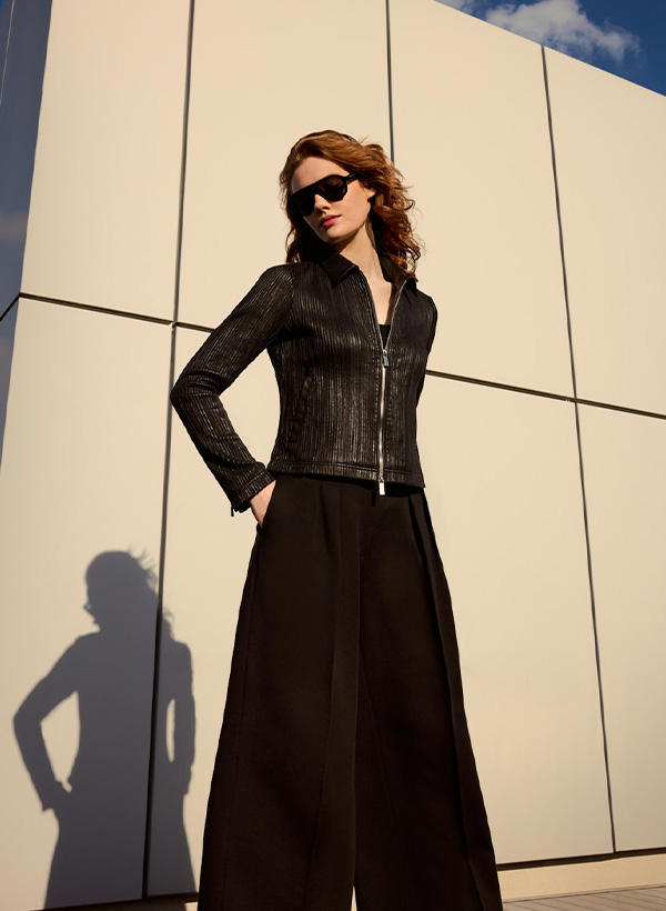 Eine Frau in einer schwarzen Lederjacke und Sonnenbrille vor einer grauen Wand in der Sonne.