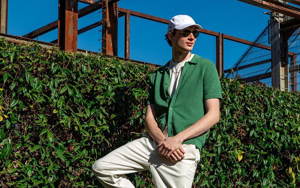 Ein Mann steht vor einer grünen Hecke, er trägt ein grünes Strickhemd, welches halb aufgeknöpft ist, und eine weiße Cap, Sonnenbrille und lässige helle Hose.