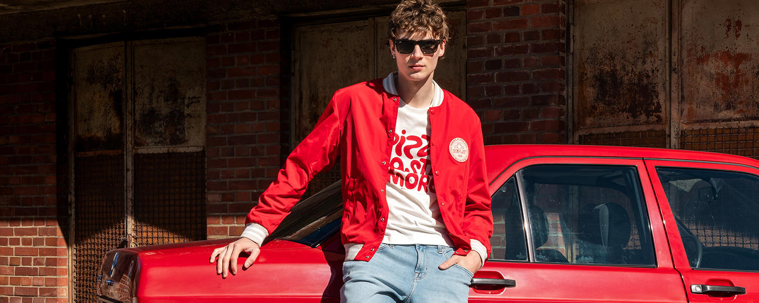 Ein junger Mann in rotem Blouson und Sonnenbrille lehnt cool an einem roten Auto