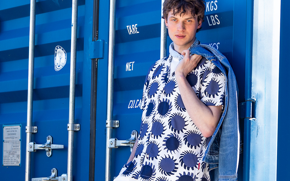 Ein Mann in einem blau-weißen Musterhemd lehnt an einem blauen Container