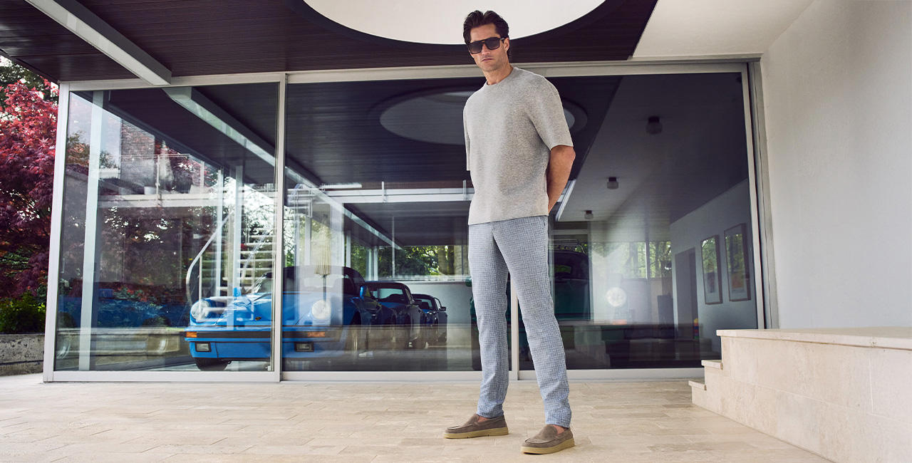 Mann in grauem Oberteil und karierten Jersey Pants von Alberto steht vor einem modernen Haus mit Glasfassade und geparktem Oldtimer im Hintergrund.