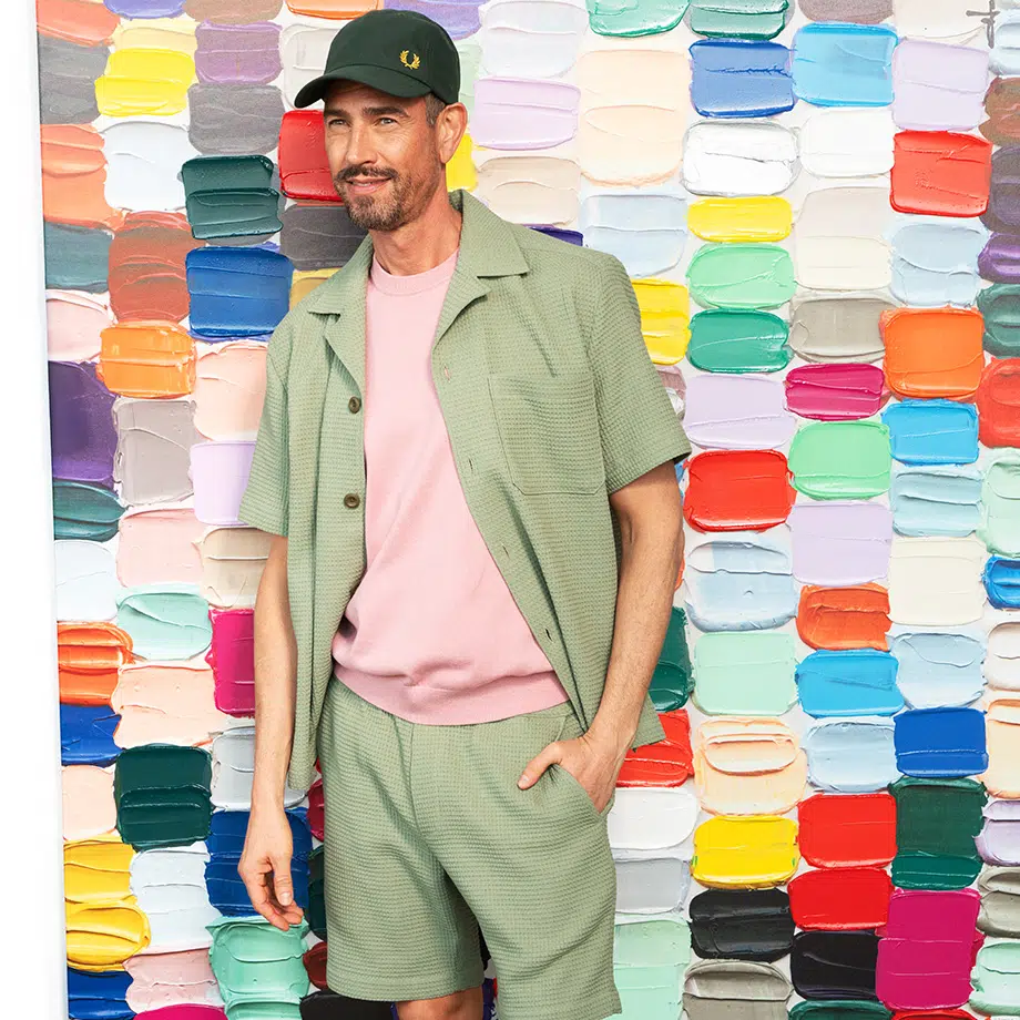 Mann mit Cap vor bunter Wand mit rechteckigen Farbflächen, trägt mintgrünes Outfit aus Hemd und Hose mit rosa T-Shirt – moderner Streetstyle-Look.