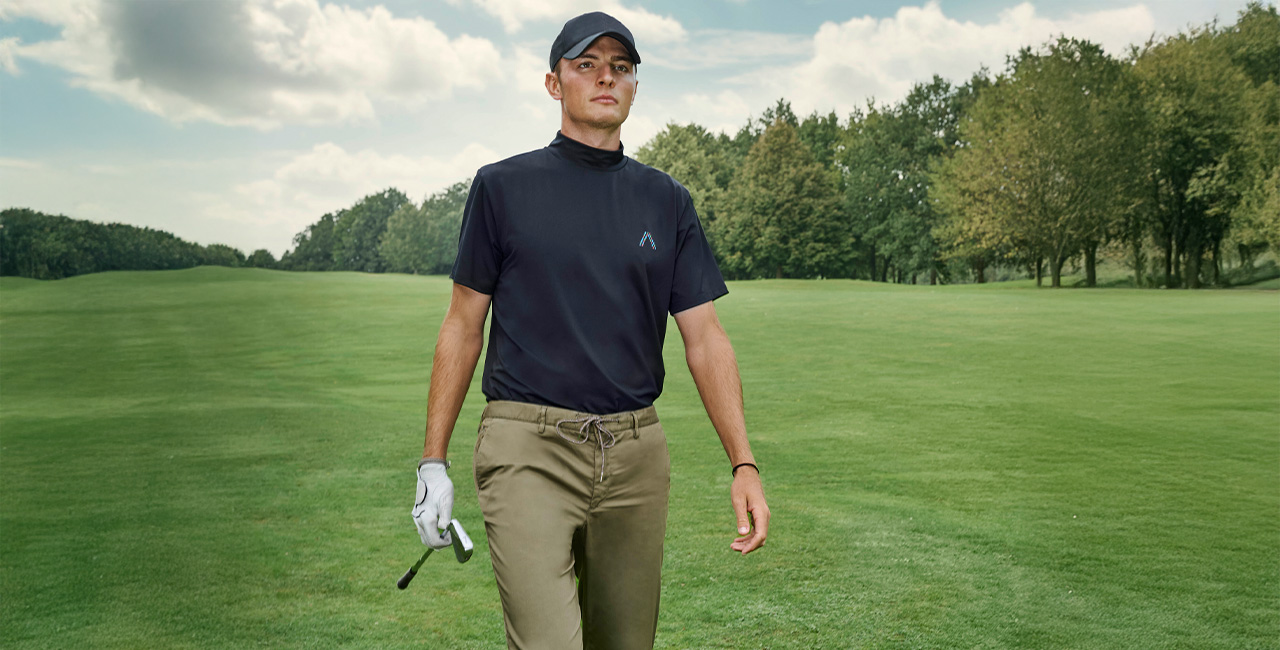Ein Mann geht auf einem Golfplatz. Er trägt ein schwarzes Polo, eine schwarze Cap und dazu passend eine grüne Golfhose.