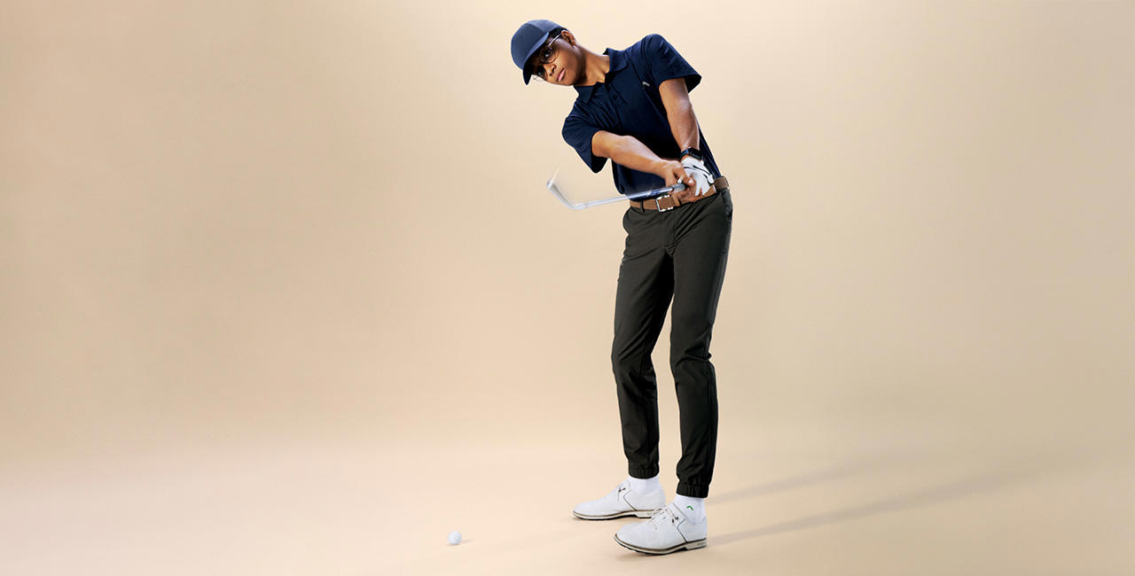 Ein junger Mann in sportlicher Pose beim Golfschwung. Er trägt ein dunkelblaues Poloshirt, eine dunkelgrüne Chinohose, weiße Sportschuhe, eine Baseballcap und eine Brille. Der Hintergrund ist einfarbig beige.