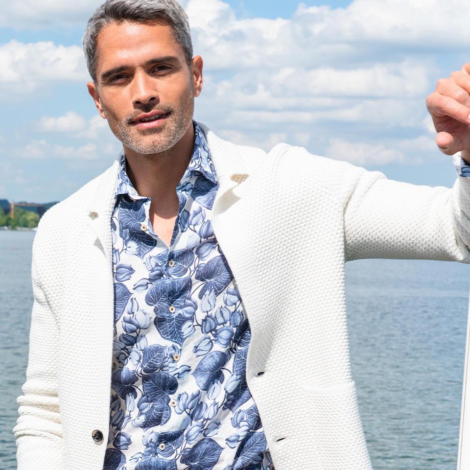 Ein Mann trägt ein weiß-blau gemustertes, florales Hemd und eine weiße Strickjacke. Im Hintergrund ist ein ruhiger See zu sehen. 