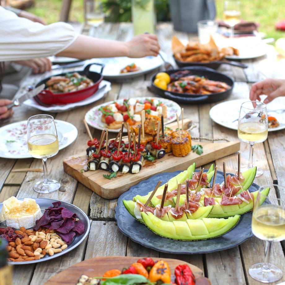 Ein reich gedeckter Holztisch im Freien mit verschiedenen Speisen, darunter Spieße mit Melone und Schinken, gefüllte Auberginenröllchen, gegrillter Mais, kleine Snacks und Wein; mehrere Hände greifen nach dem Essen.