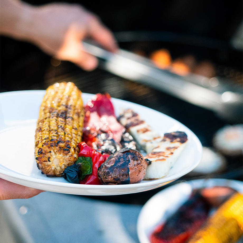 Ein weißer Teller mit gegrilltem Gemüse, darunter ein gegrillter Maiskolben, Paprika, Halloumi und eine kleine Kartoffel, im Vordergrund; im Hintergrund ein Grill und eine Hand mit einer Grillzange.