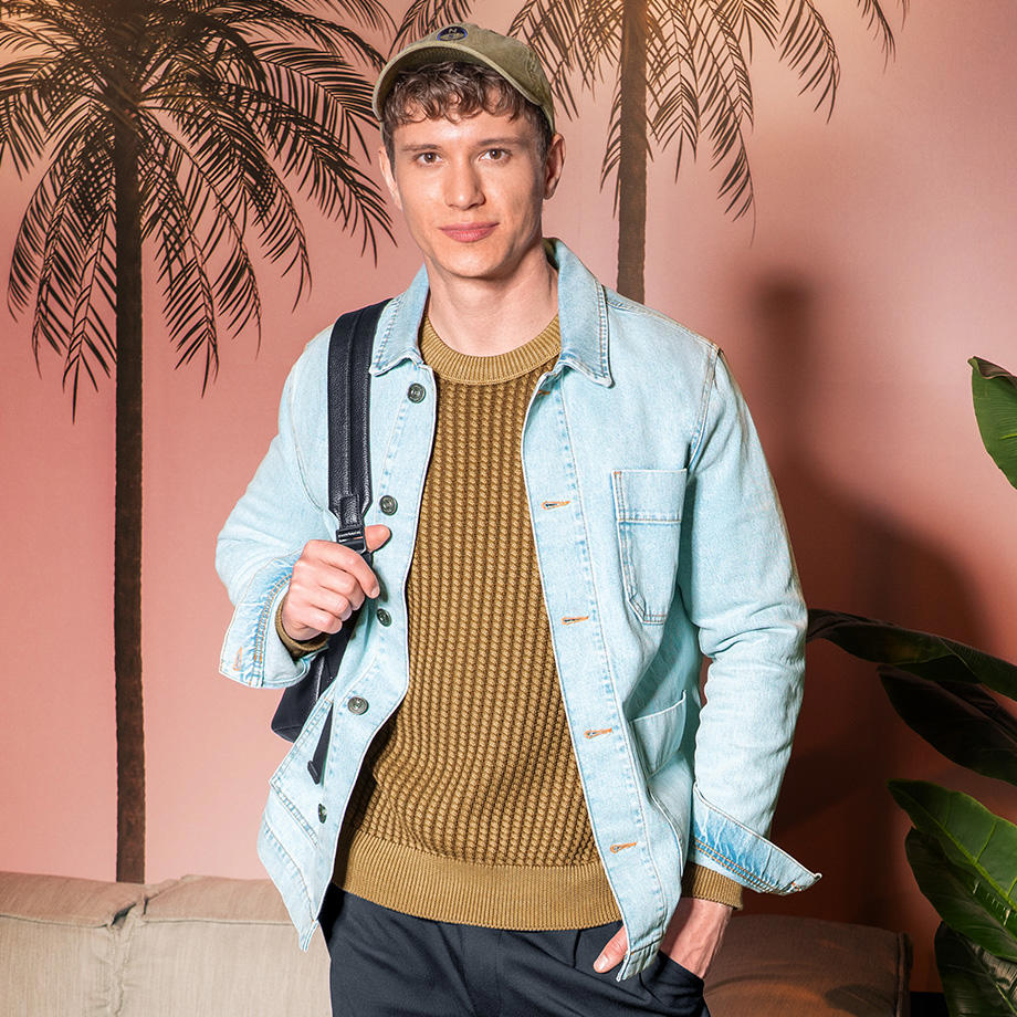 Ein junger Mann steht vor einer Wand mit Palmenmotiv. Er trägt eine hellblaue Jeansjacke über einem olivgrünen Strickpullover, eine dunkelblaue Hose und eine beigefarbene Baseballkappe. Über der Schulter trägt er einen schwarzen Rucksack.