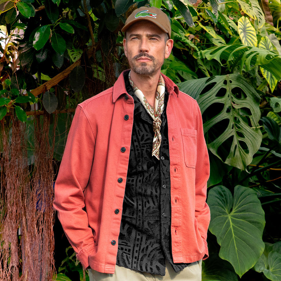 Ein Mann steht vor einer üppigen grünen Pflanzenkulisse. Er trägt eine korallenrotes Overshirt über einem schwarzen Hemd mit Muster und eine beige Hose. Abgerundet wird das Outfit durch ein Nickituch und eine Baseballkappe.