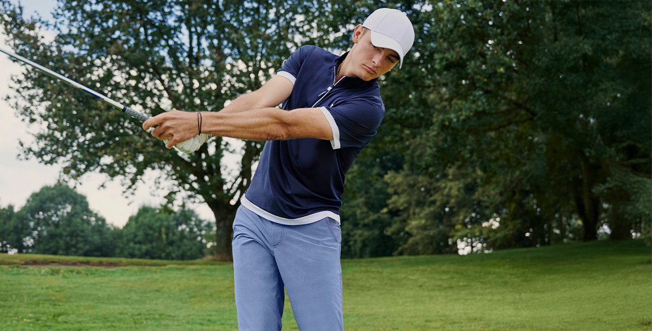 Ein junger Mann in sportlicher Golfbekleidung schlägt einen Golfschläger auf einem gepflegten Golfplatz. Er trägt ein dunkelblaues Poloshirt mit weißen Akzenten, eine weiße Baseballkappe und hellblaue Hosen.