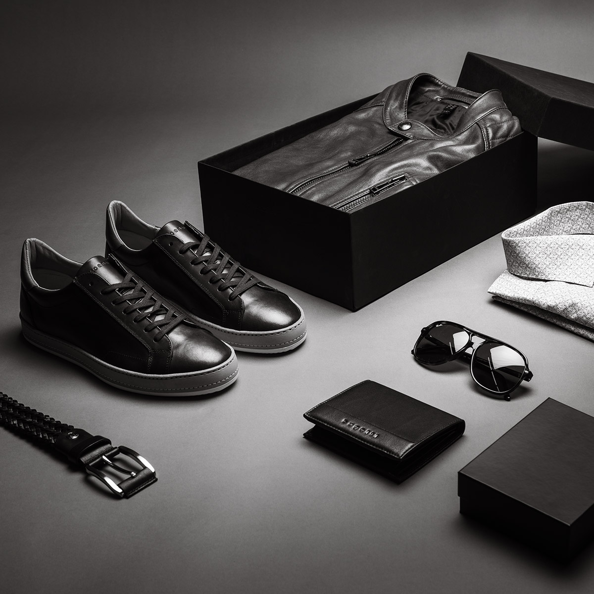 Ein Flatlay einer Gruppe von Produkten: Ledersneaker, Lederjacke, Sonnenbrille, zwei Ledergürtel, Businesshemd und Lederportemonnaie. Es ist in schwarz-weiß.