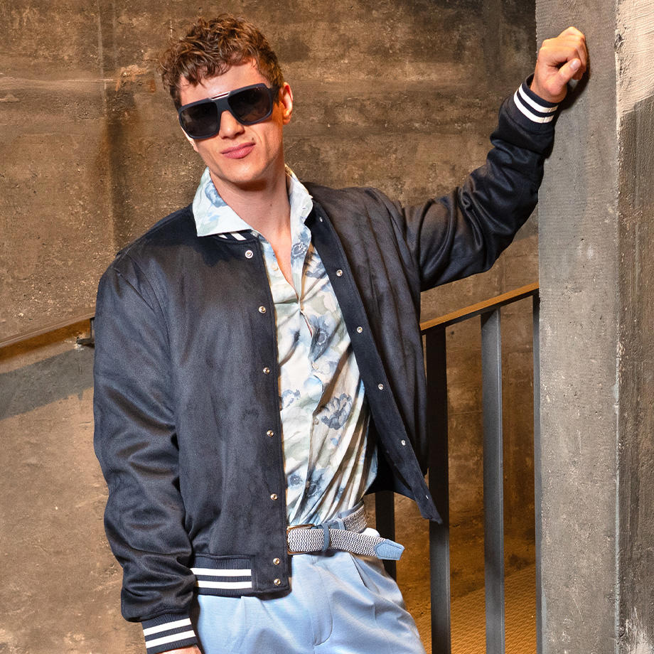 Ein junger Mann lehnt lässig an einer Betonwand, trägt eine dunkle Collegejacke, ein gemustertes Hemd, helle Hose und große Sonnenbrille.