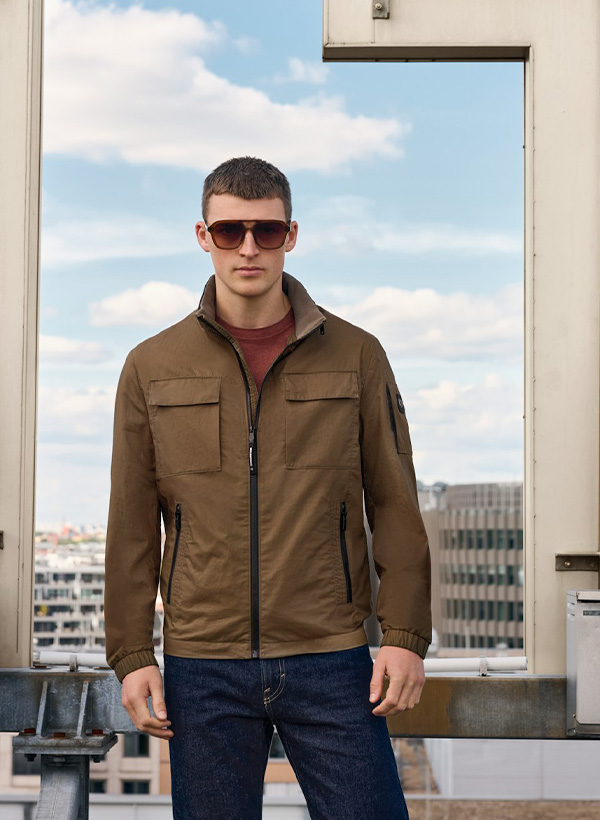 Ein junger Mann trägt eine braune Funktionsjacke und eine Sonnenbrille. Er steht auf einem Dach in einer Großstadt.