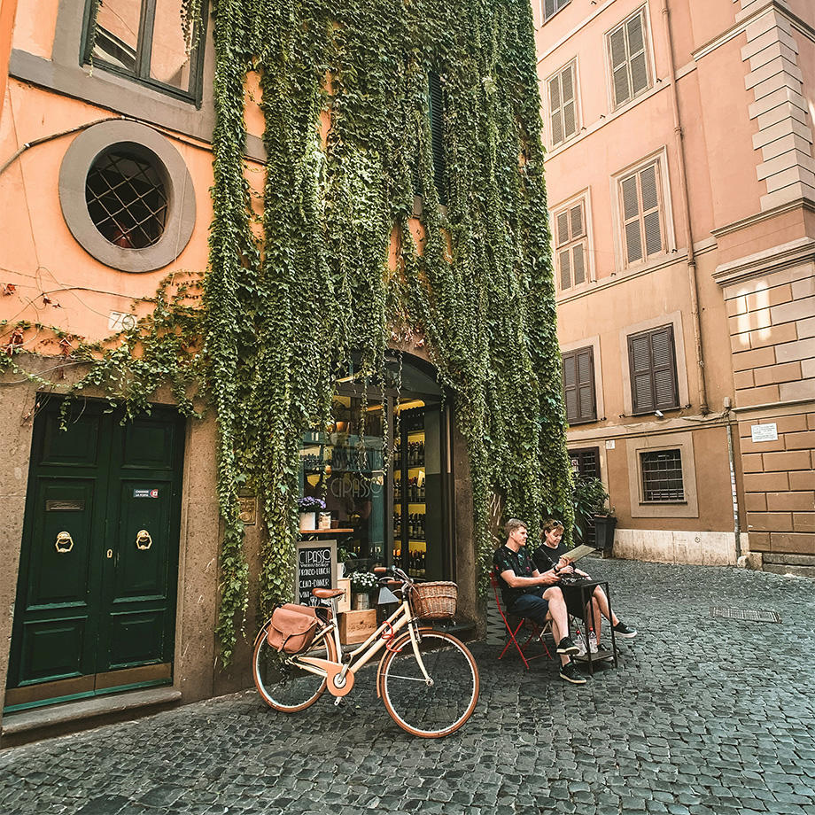 Ein Foto einer Gasse in einer Stadt. An einem Haus hängen Ranken von Efeu und unten ist ein Schaufenster und Eingang zu einem kleinen Cafe oder Restaurant. Davor steht ein Retro-Citybike und zwei Menschen sitzen an einem kleinen Tisch.