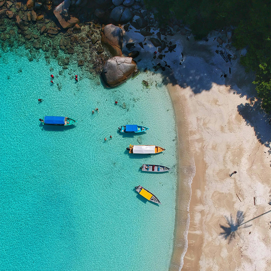 Ein Drohnenfoto eines Strands von oben. Türkises Wasser, Schatten von Palmen und einige kleine Fischerboote.
